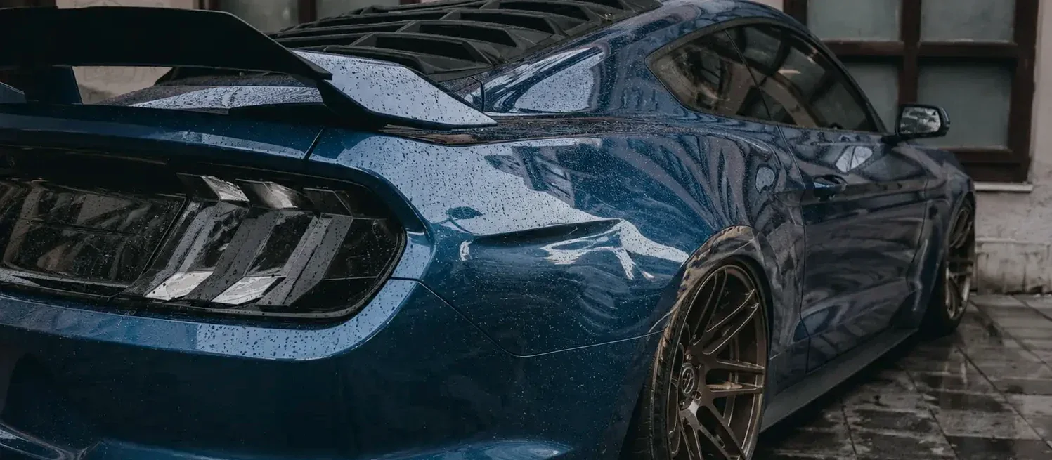 Rain beaded on the rear quarter of a blue Ford Mustang parked on wet pavers, bronze wheels catching what light there is.