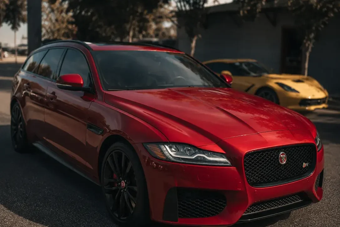 A red Jaguar XF Sportbrake on black wheels parked on a sunlit lot, a yellow Corvette behind it.